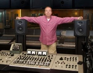 Geoff Emerick standing with his arms outstretched behind a large studio mixing console between two monitor speakers.