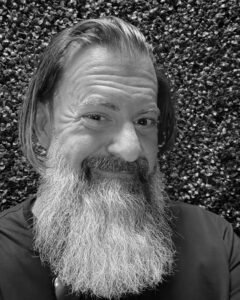 Loren Weisman, brand communications and optics speaker, facing the camera with a full grey beard and shoulder-length hair, wearing a maroon shirt in front of a green hedge wall. 8x10 black and white promo photo of brand communications and optics speaker Loren Weisman.