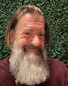 Loren Weisman, brand communications and optics speaker, facing the camera with a full grey beard and shoulder-length hair, wearing a maroon shirt in front of a green hedge wall. 8x10 colour promo photo of brand communications and optics speaker Loren Weisman.