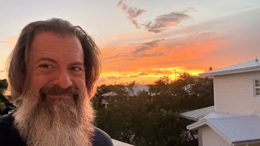 Loren Weisman outdoors at sunset, smiling, with an orange sky and treeline behind him.
