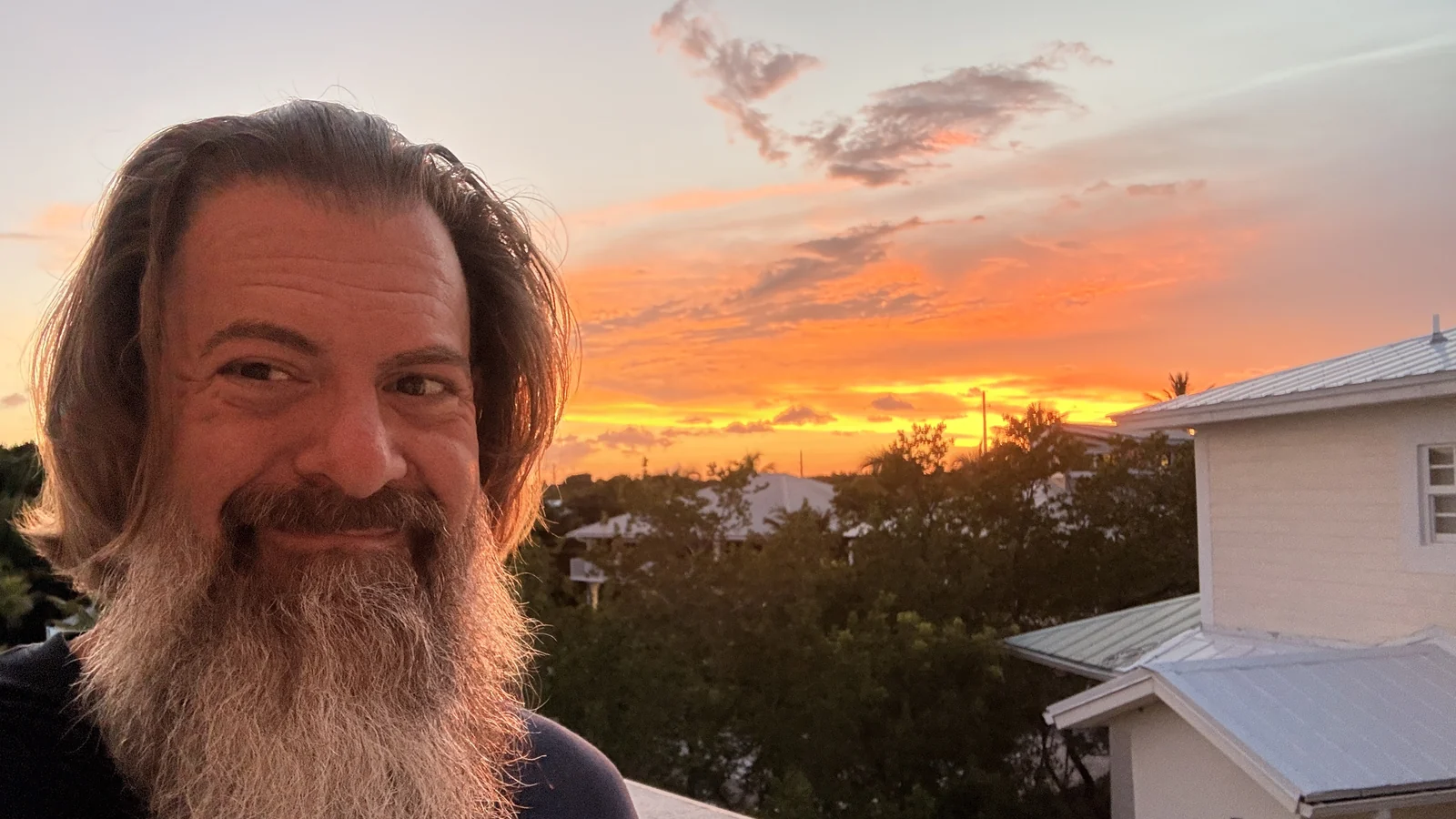 Loren Weisman outdoors at sunset, smiling, with an orange sky and treeline behind him.