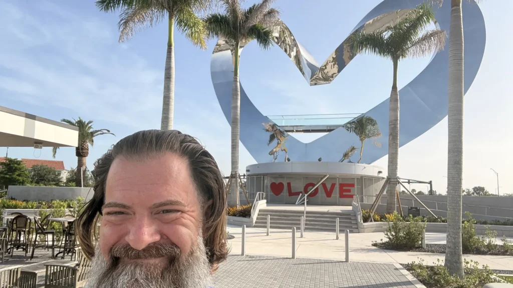 Loren Weisman outdoors beside a public art sculpture, palm trees and clear sky behind him.