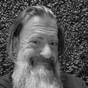 Loren Weisman, brand communications and optics speaker, facing the camera with a full grey beard and shoulder-length hair, wearing a maroon shirt in front of a green hedge wall. Square black and white headshot of brand communications and optics speaker Loren Weisman.
