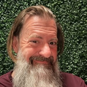 Loren Weisman, brand communications and optics speaker, facing the camera with a full grey beard and shoulder-length hair, wearing a maroon shirt in front of a green hedge wall. Square colour headshot of brand communications and optics speaker Loren Weisman.