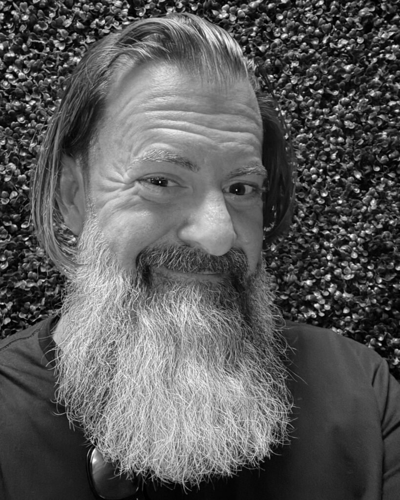 Loren Weisman, brand communications and optics speaker, facing the camera with a full grey beard and shoulder-length hair, in front of a hedge wall. Black and white. Headshot.