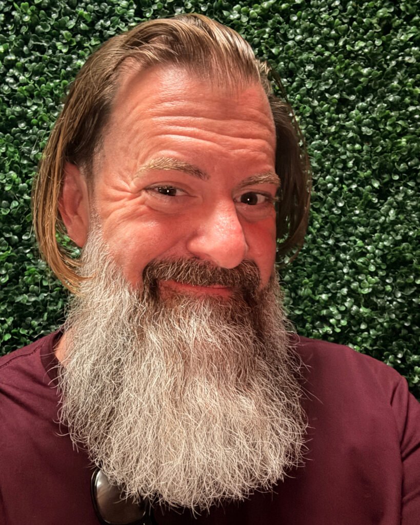 Loren Weisman, brand communications and optics speaker, facing the camera with a full grey beard and shoulder-length hair, wearing a maroon shirt in front of a green hedge wall. 8x10 headshot, color.