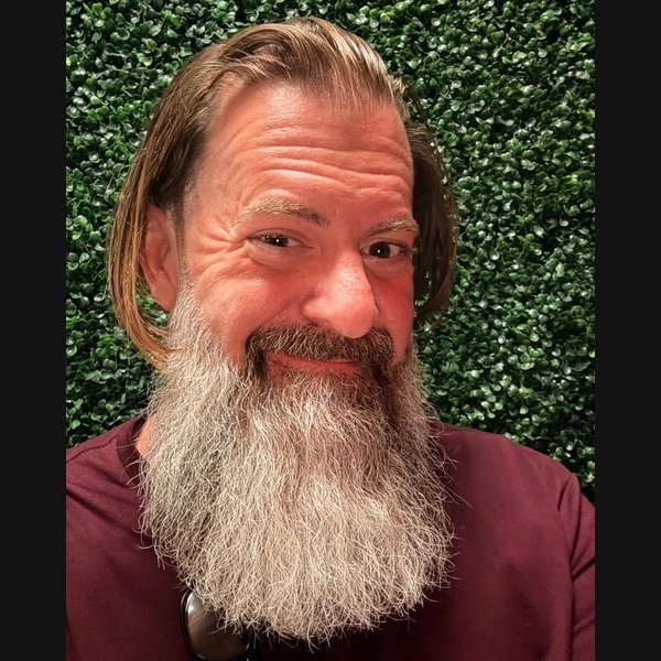 Loren Weisman smiling, with long hair, a full gray beard, and a maroon shirt